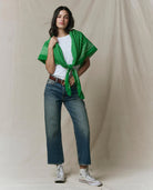 Woman wearing a green patterned shawl over a white shirt and blue jeans on a beige background