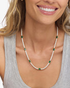 A close-up lifestyle shot of a woman wearing a delicate necklace. The necklace features a dual-strand design with a line of small white pearls and a line of clear round crystals, accented by small green oval stones and larger green oval center stones set in a silver-toned metal.