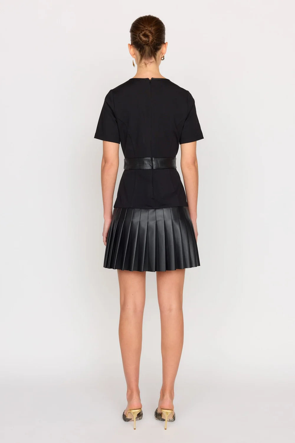 A model stands facing backwards against a light gray studio background wearing a black short-sleeve top with a matching black pleated mini skirt and a thin black waist belt.