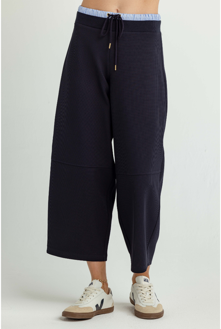 A person wearing dark navy wide-leg cropped sweatpants with a drawstring waist and light blue striped elastic band, paired with white sneakers, against a light gray studio background.