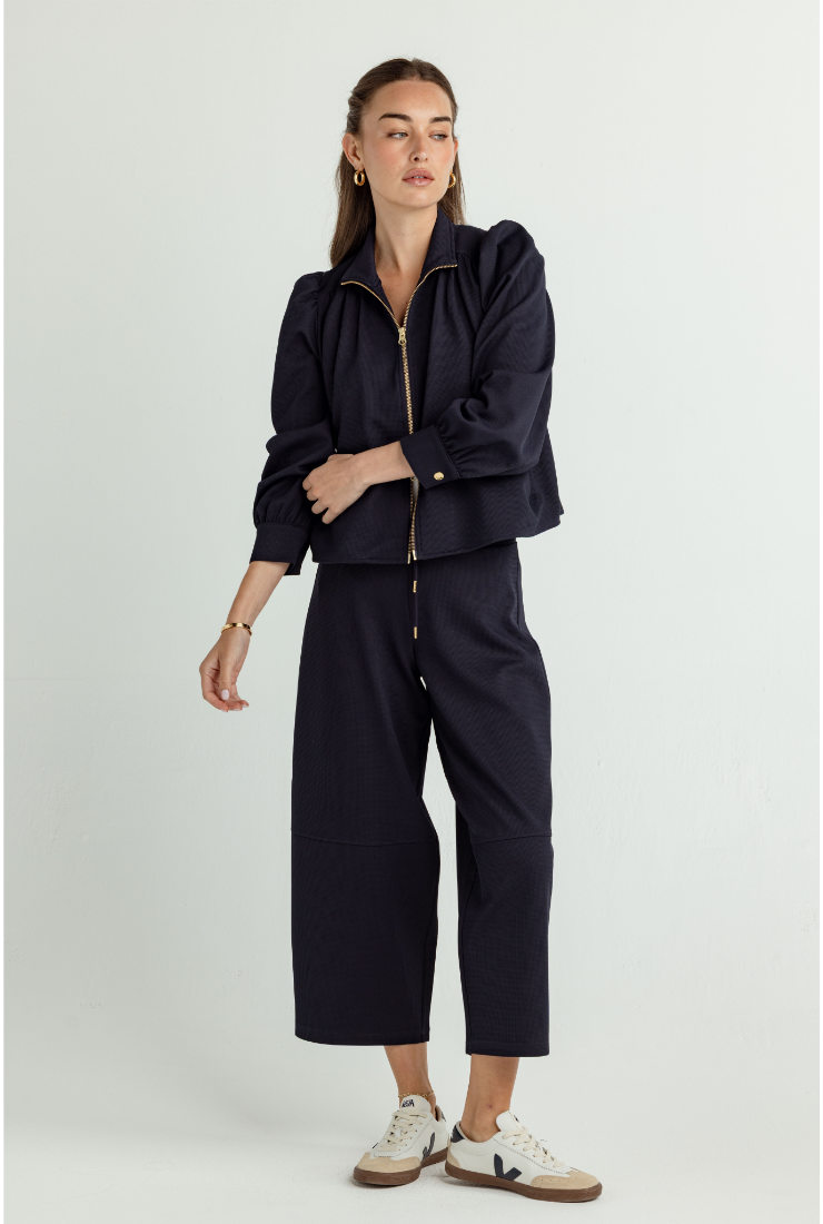 A model stands against a light gray studio background wearing a dark navy two-piece set consisting of a cropped zip-up jacket with puffed sleeves and matching cropped pants, paired with white sneakers.