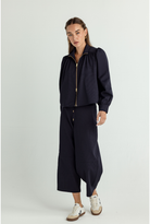 A woman modeling a dark navy blue two-piece outfit consisting of a cropped long-sleeve zip-up jacket with gold-tone zipper details and matching wide-leg cropped pants, paired with white sneakers, against a light gray studio background.