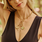 A close-up lifestyle shot of a model wearing a gold-toned chain necklace with three dangling charms: a horseshoe, a bow, and a round medallion, paired with a black V-neck top.