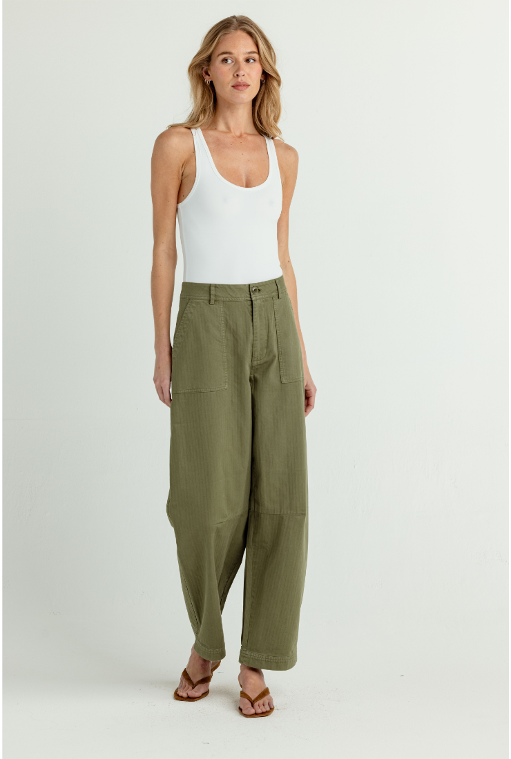 A woman modeling a pair of high-waisted, wide-leg olive green pants with a white tank top and brown sandals, standing against a plain light gray studio background.
