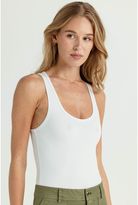 A woman wearing a white scoop-neck tank top and olive green pants, photographed against a clean light gray studio background.