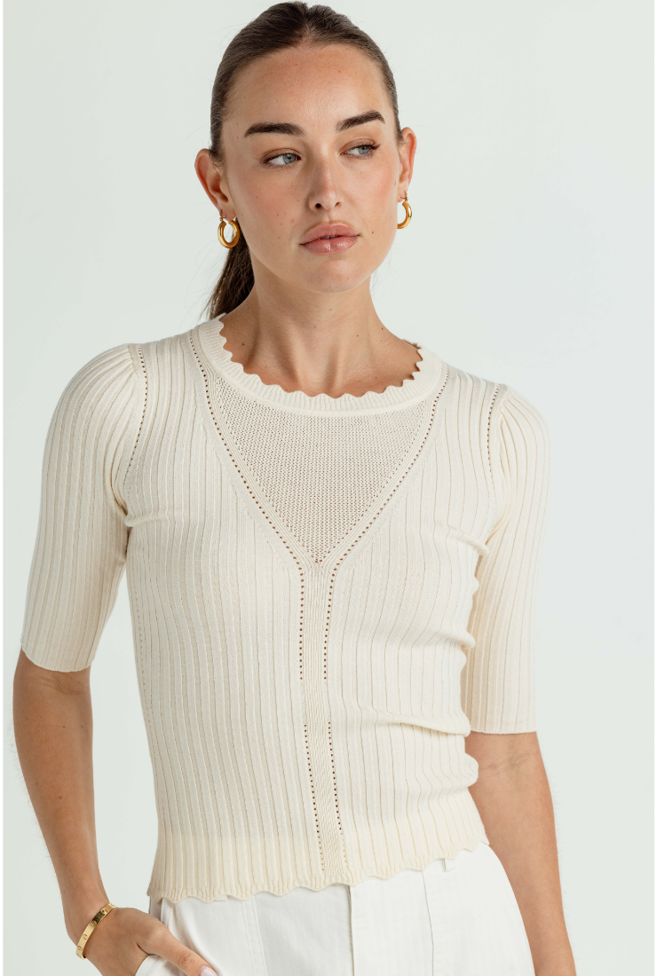 A woman wearing a cream-colored short-sleeve ribbed knit top with a scalloped neckline and a V-shaped openwork chest detail, paired with white trousers, against a light gray studio background.