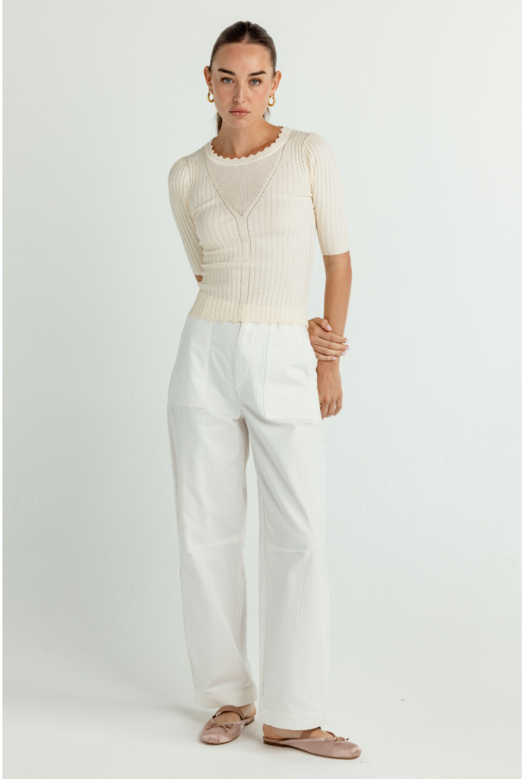 A woman modeling a cream ribbed short-sleeve knit top with a scalloped neckline and white wide-leg trousers, standing against a light gray studio background.