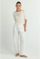 A woman modeling a cream ribbed short-sleeve knit top with a scalloped neckline and white wide-leg trousers, standing against a light gray studio background.