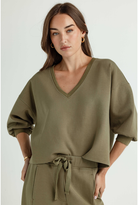 A woman wearing an olive green V-neck sweatshirt with voluminous sleeves and a matching drawstring waist pants, photographed against a light gray studio background.
