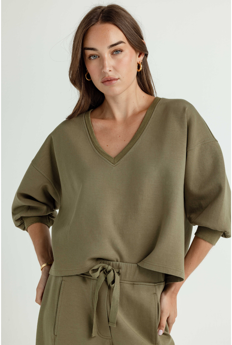 A woman wearing an olive green V-neck sweatshirt with voluminous sleeves and a matching drawstring waist pants, photographed against a light gray studio background.