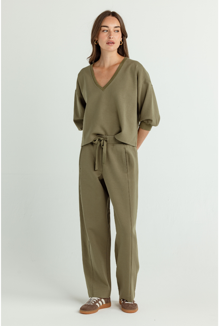 A woman modeling a matching olive green lounge set consisting of a short-sleeve V-neck top with a drawstring waist and relaxed-fit pants, paired with brown sneakers, against a plain light gray studio background.