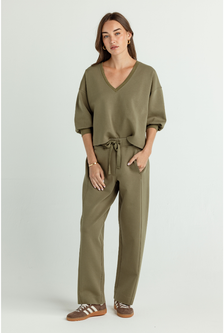 A woman modeling an olive green lounge set consisting of a V-neck top with elbow-length sleeves and matching wide-leg drawstring pants, paired with brown sandals, against a light gray studio background.