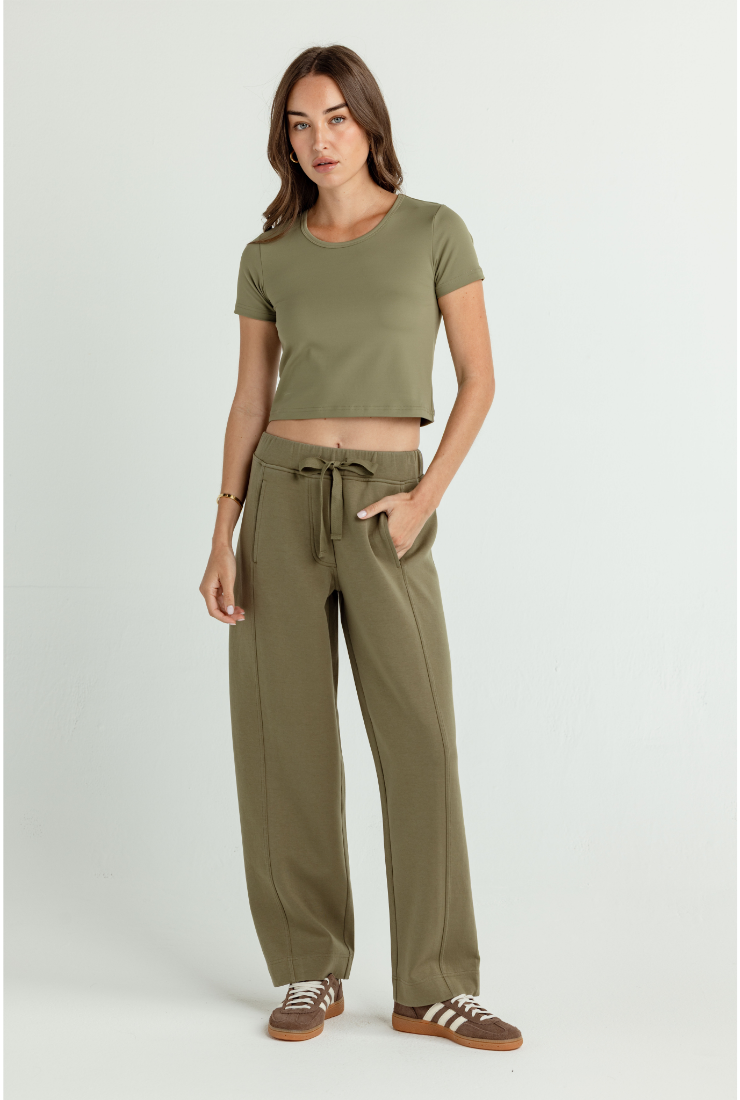 A woman modeling an olive green two-piece lounge set consisting of a short-sleeve crop top and matching wide-leg drawstring pants, paired with brown and white sneakers, against a light gray studio background.