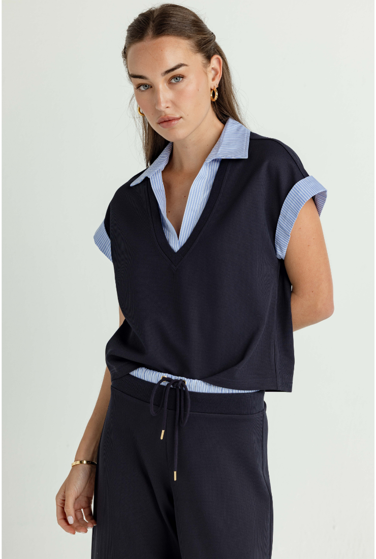A woman wearing a navy sleeveless knit vest over a light blue and white striped collared shirt, paired with navy drawstring trousers, against a plain light gray studio background.