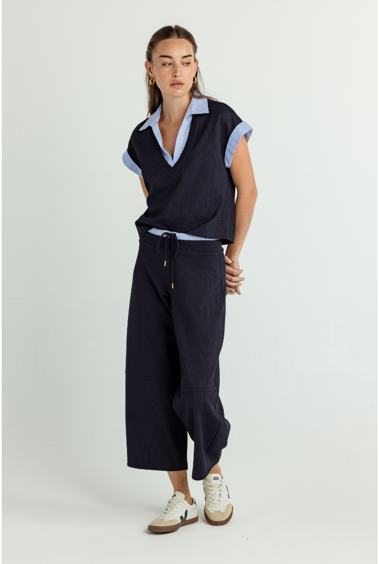 A woman modeling a dark navy sleeveless polo shirt vest over a light blue collared shirt, paired with dark navy wide-leg cropped pants and white sneakers, against a light gray studio background.