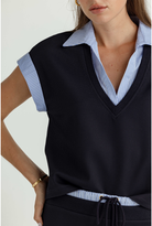 A close-up of a woman wearing a dark navy ribbed knit V-neck sleeveless top over a light blue and white striped collared shirt, with a matching navy drawstring waist detail visible at the bottom.