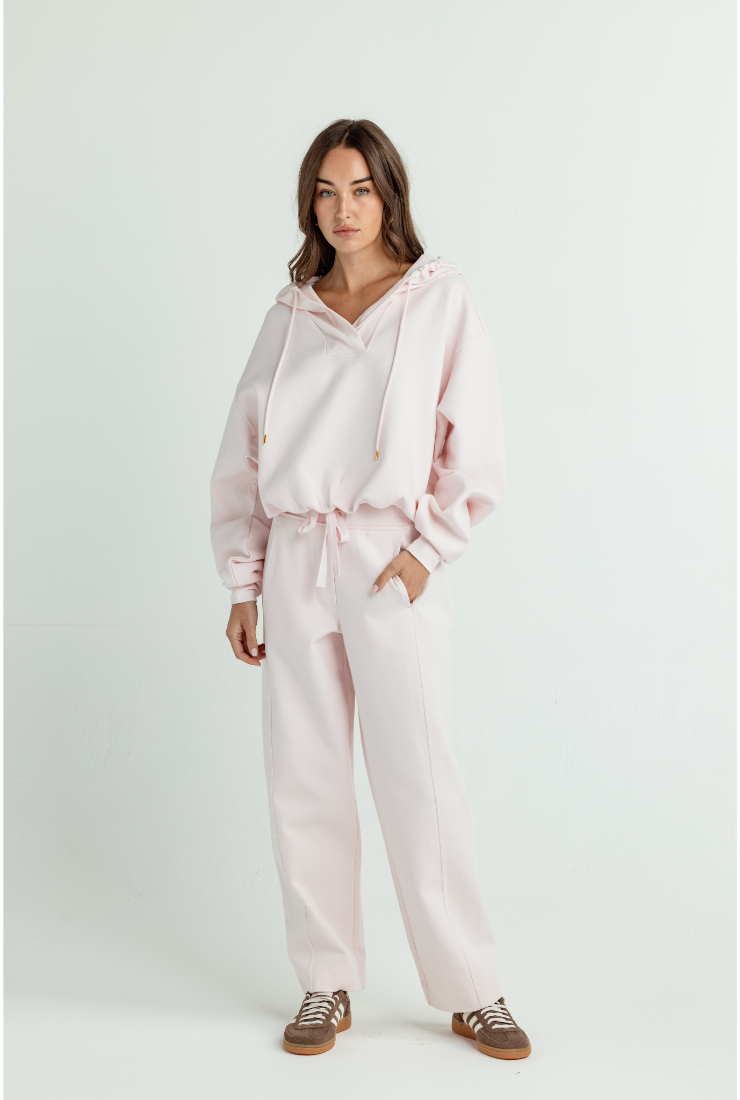 A woman modeling a light pink two-piece loungewear set consisting of a cropped drawstring hoodie and matching wide-leg sweatpants, paired with brown sneakers, against a plain light gray studio background.