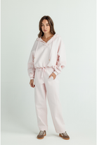 A woman modeling a light pink two-piece loungewear set consisting of a cropped drawstring hoodie and matching wide-leg sweatpants, paired with brown sneakers, against a plain light gray studio background.