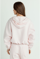 A woman shown from the back wearing a light pink cropped hoodie with a ruffled hood trim and matching light pink sweatpants against a plain light background.