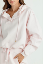 A close-up of a woman wearing a light pink hooded sweatshirt and matching drawstring pants against a plain light background.