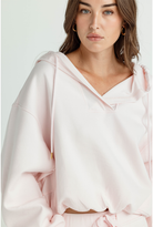 A woman wearing a light pink oversized hooded sweatshirt with drawstrings and a V-neckline, shown in a close-up studio shot against a plain light background.