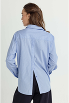A woman shown from the back wearing a light blue and white vertically striped button-down shirt with a relaxed fit and black trousers, photographed against a plain light background.