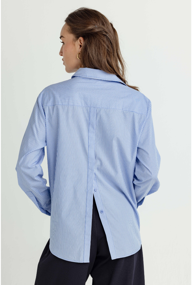 A woman shown from the back wearing a light blue and white vertically striped button-down shirt with a relaxed fit and black trousers, photographed against a plain light background.