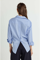 A woman shown from the back wearing a light blue and white vertically striped button-down shirt with rolled-up sleeves and dark trousers, against a plain light background.
