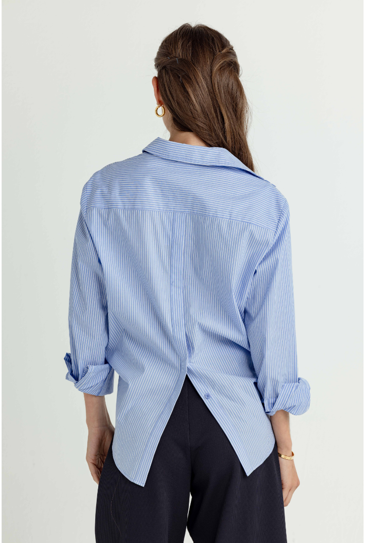 A woman shown from the back wearing a light blue and white vertically striped button-down shirt with rolled-up sleeves and dark trousers, against a plain light background.