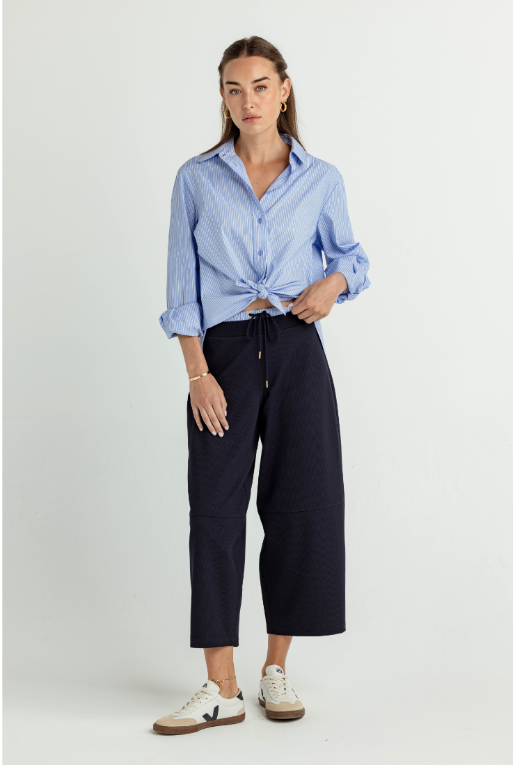 A woman modeling a light blue long-sleeve button-up shirt tied at the waist with dark navy cropped trousers and white sneakers, photographed against a plain light gray studio background.
