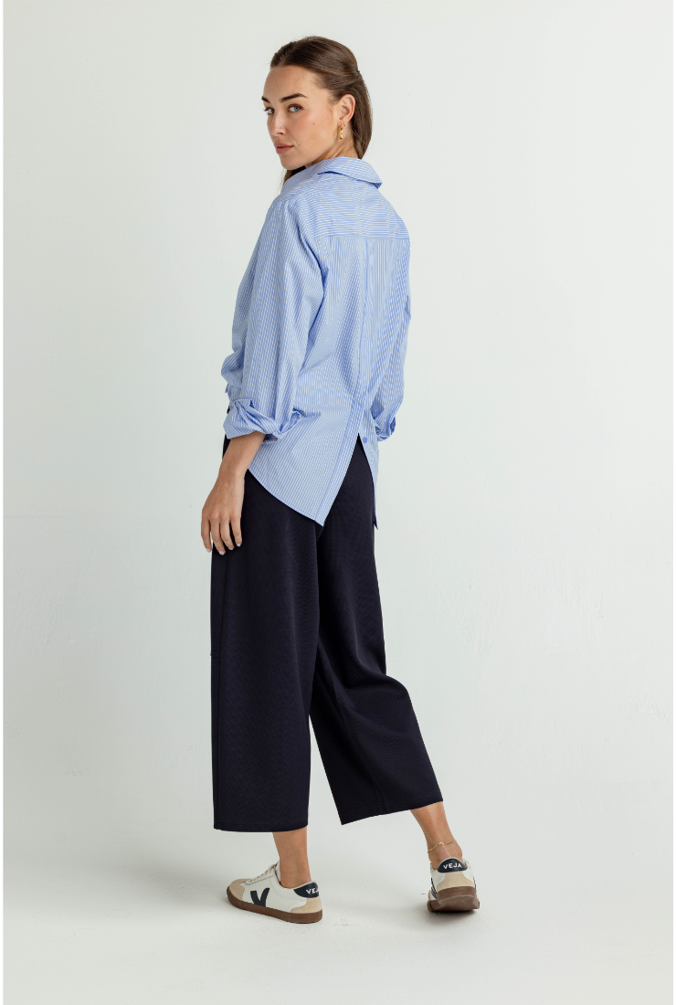 A woman modeling a light blue long-sleeve button-up shirt with rolled cuffs and dark navy cropped wide-leg pants, standing against a plain light gray studio background.