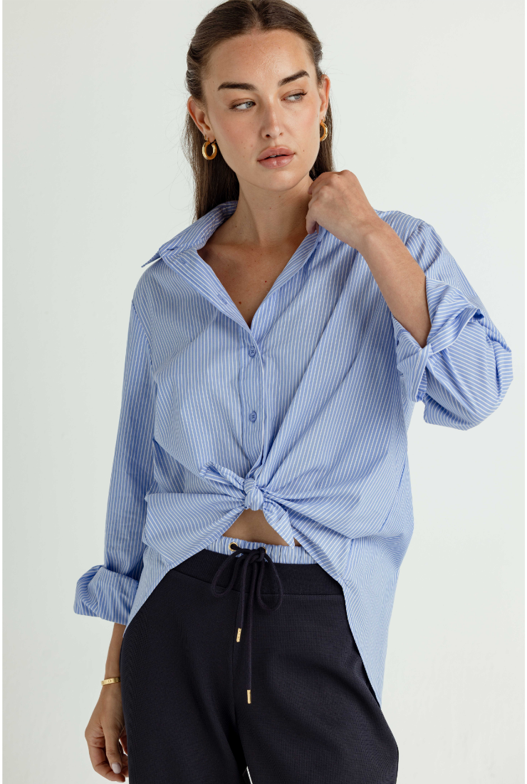 A woman wearing a light blue and white striped button-down shirt tied at the waist, paired with dark drawstring trousers, against a plain light background.