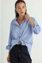 A woman wearing a light blue and white striped button-down shirt tied at the waist, paired with dark drawstring trousers, against a plain light background.
