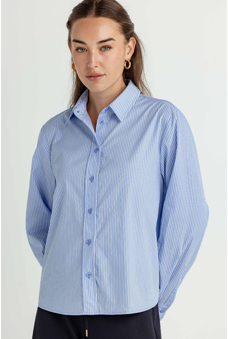 A woman wearing a light blue and white vertically striped button-down shirt, paired with dark pants, photographed against a plain light background.
