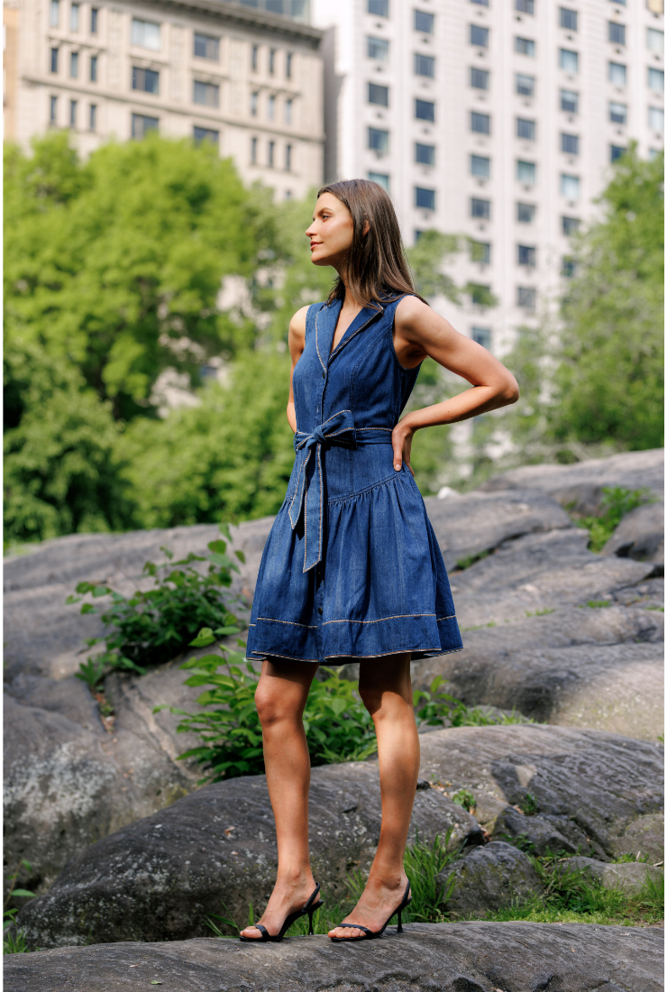 A woman wearing a sleeveless blue denim dress with a tie waist and tiered skirt, standing outdoors on large rocks with trees and buildings in the background.