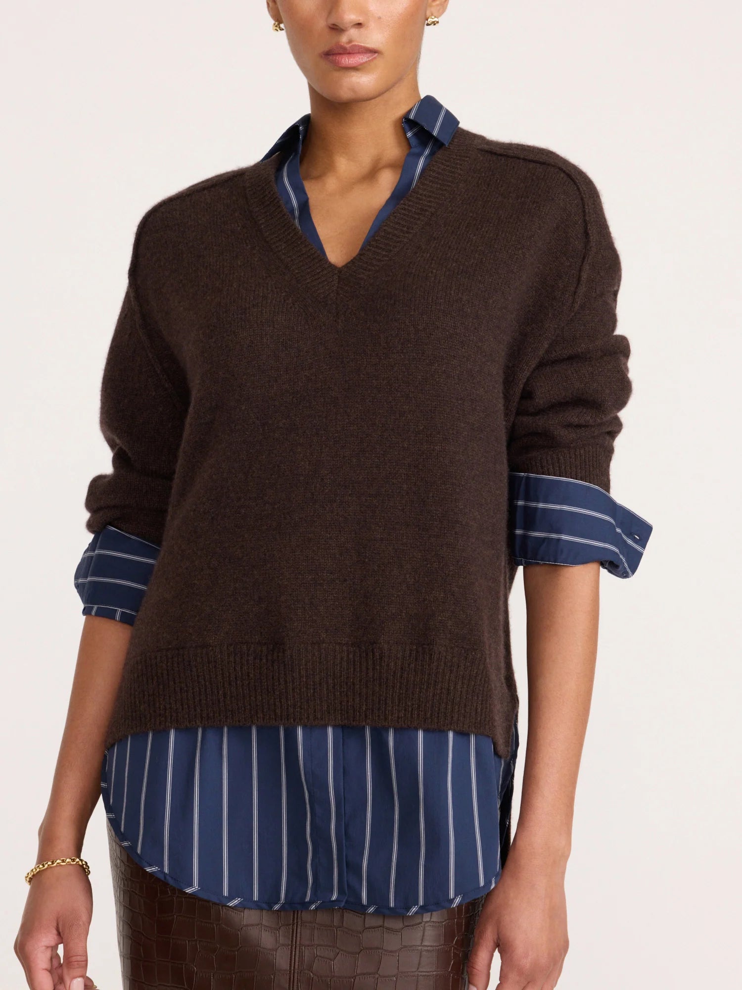 A model wearing a dark brown V-neck knit sweater layered over a blue and white striped button-down shirt, shown from the neck down against a plain light background.