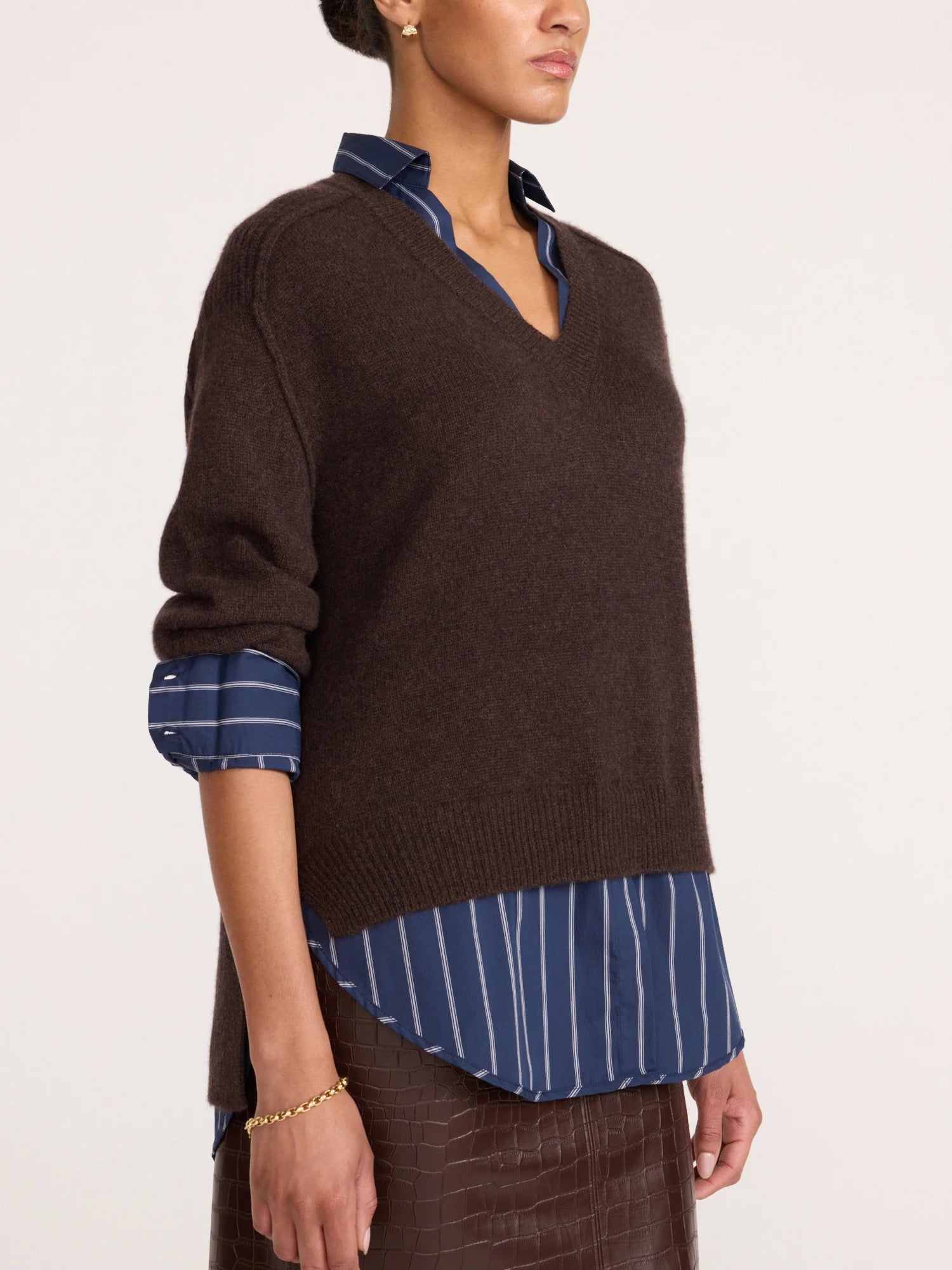 A model wearing a dark brown V-neck knit sweater layered over a blue and white striped button-down shirt, shown from the chest down against a plain light background.
