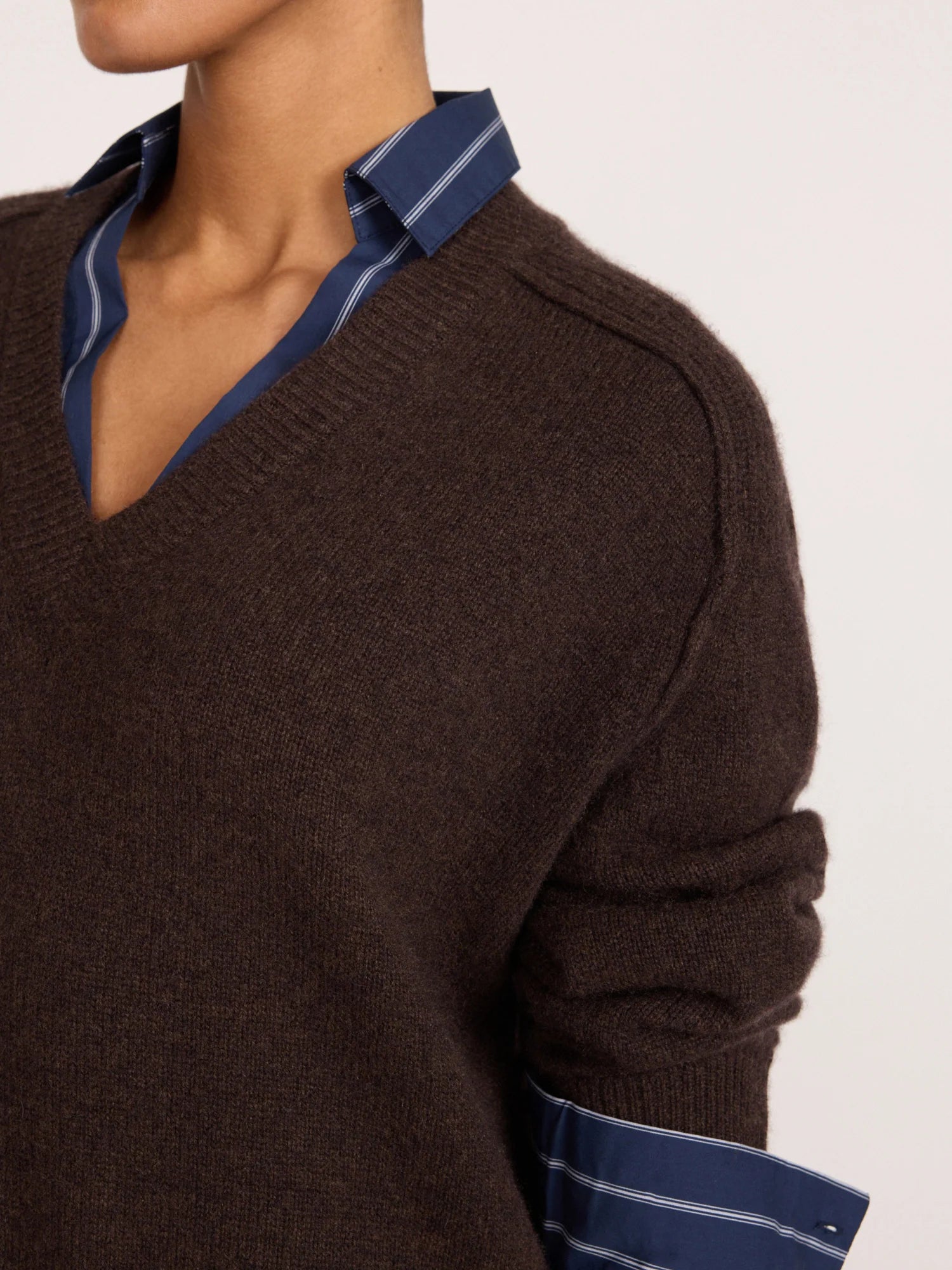 A close-up of a person wearing a dark brown V-neck knit sweater layered over a blue striped dress shirt, shown against a plain light background.
