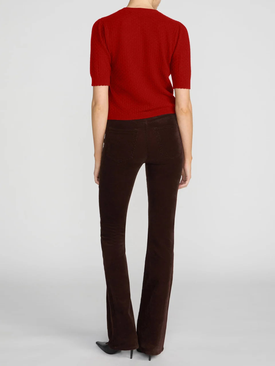 A model shown from the back wearing a short-sleeve red knit top and dark brown flared trousers, paired with black pointed-toe heels, against a light gray studio background.