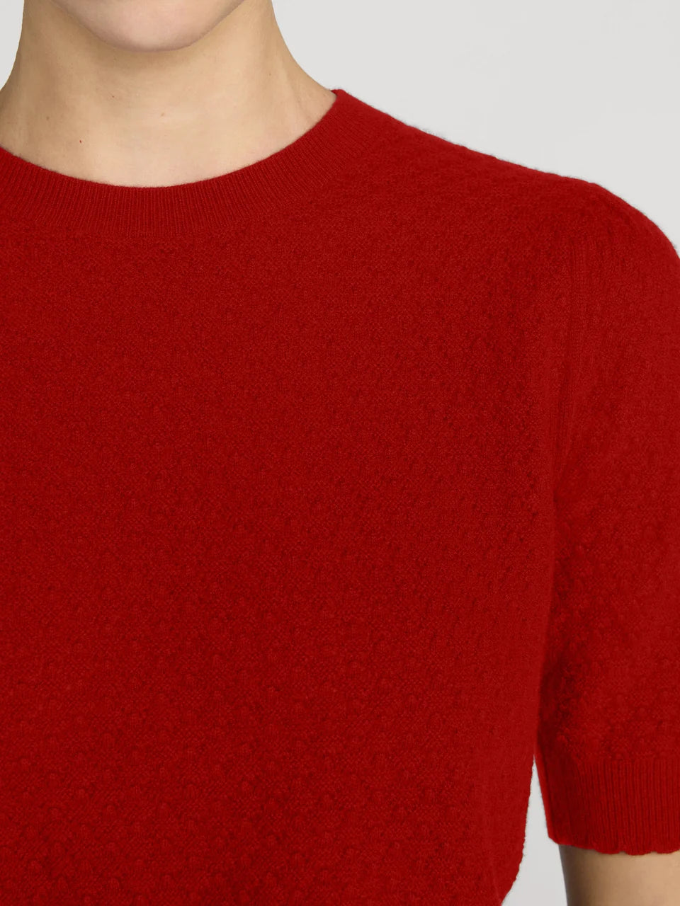 A close-up of a person wearing a bright red short-sleeve knit sweater against a light gray background.