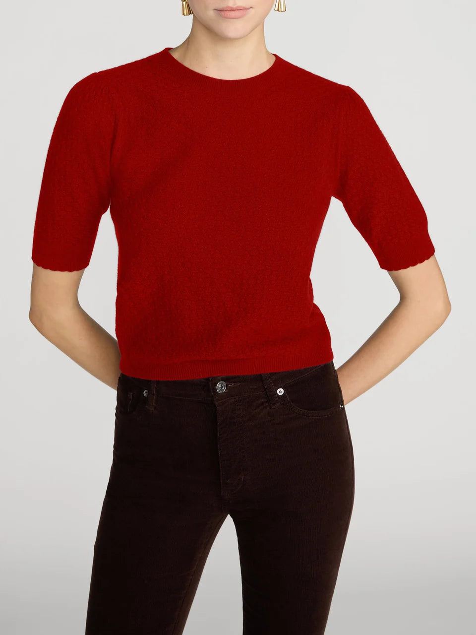 A woman wearing a bright red short-sleeve knit top and dark brown pants, cropped from the mouth down against a light gray studio background.