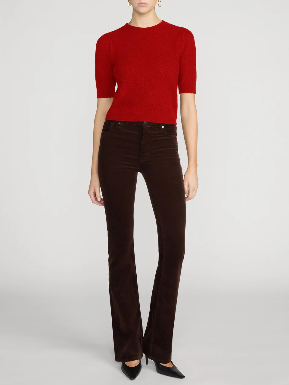 A model wearing a bright red short-sleeve knit top and dark brown velvet flare pants, paired with black pointed-toe heels, against a light gray studio background.