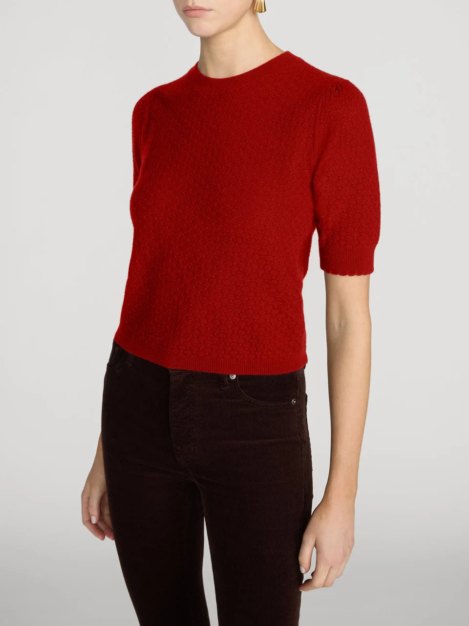 A model wearing a textured red short-sleeve knit sweater and dark brown pants, shown from the chin down against a light gray studio background.