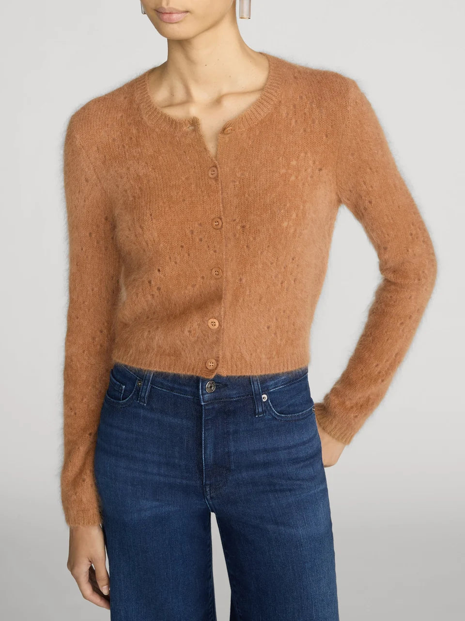 A model wearing a fuzzy, cropped button-front cardigan in a warm camel brown color, paired with dark blue jeans, against a light gray studio background.