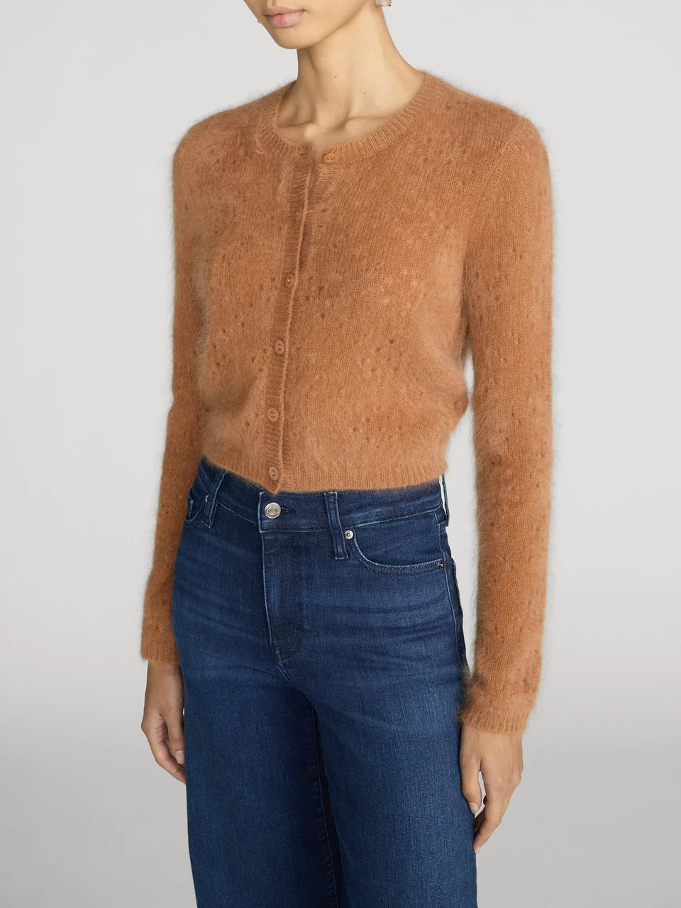 A woman wearing a fuzzy, cropped button-front cardigan in a warm camel brown color, paired with dark blue jeans, against a light gray studio background.