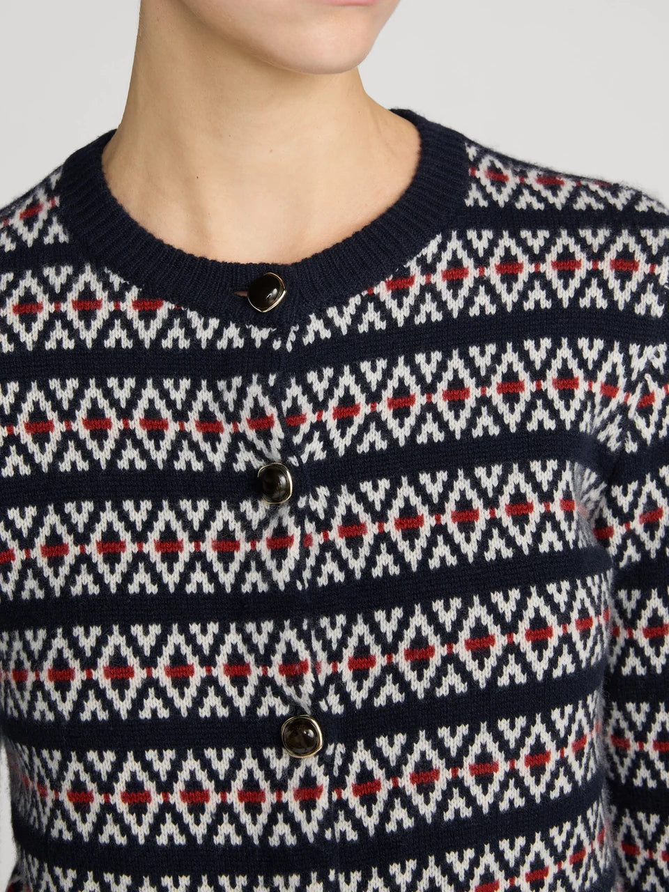 Close-up of a person wearing a navy knit cardigan sweater with a geometric fair isle pattern in white and red, featuring dark buttons on a light gray background.