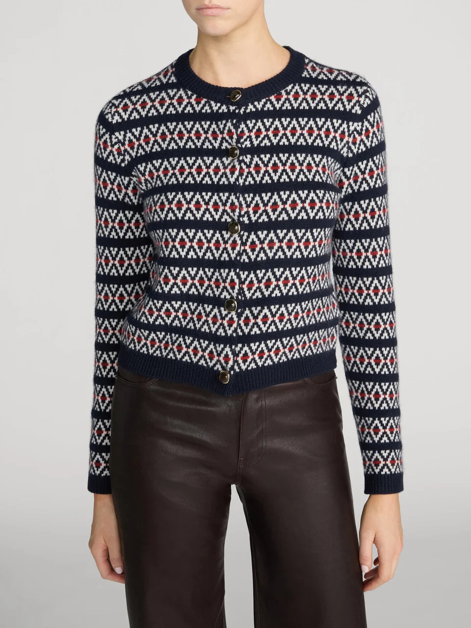 A model wearing a navy blue cardigan with a red and white geometric Fair Isle pattern, paired with dark brown leather pants, against a light gray studio background.