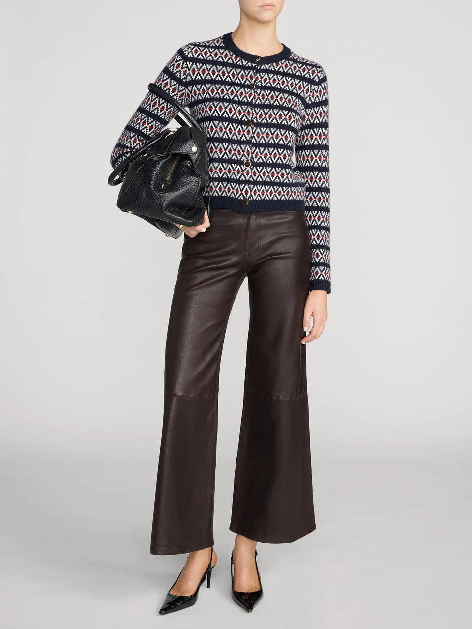 A model wearing a navy, beige, and white patterned knit sweater, dark brown wide-leg leather pants, and black pointed-toe heels, holding a black handbag against a light gray studio background.