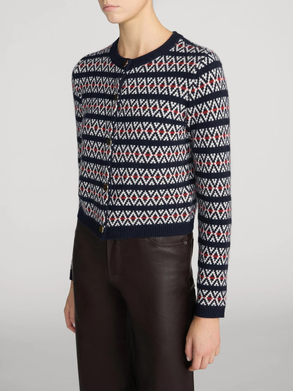 A cropped knit cardigan sweater with a navy blue base and a red and white geometric fair isle pattern, worn by a model with dark brown leather pants against a light gray studio background.