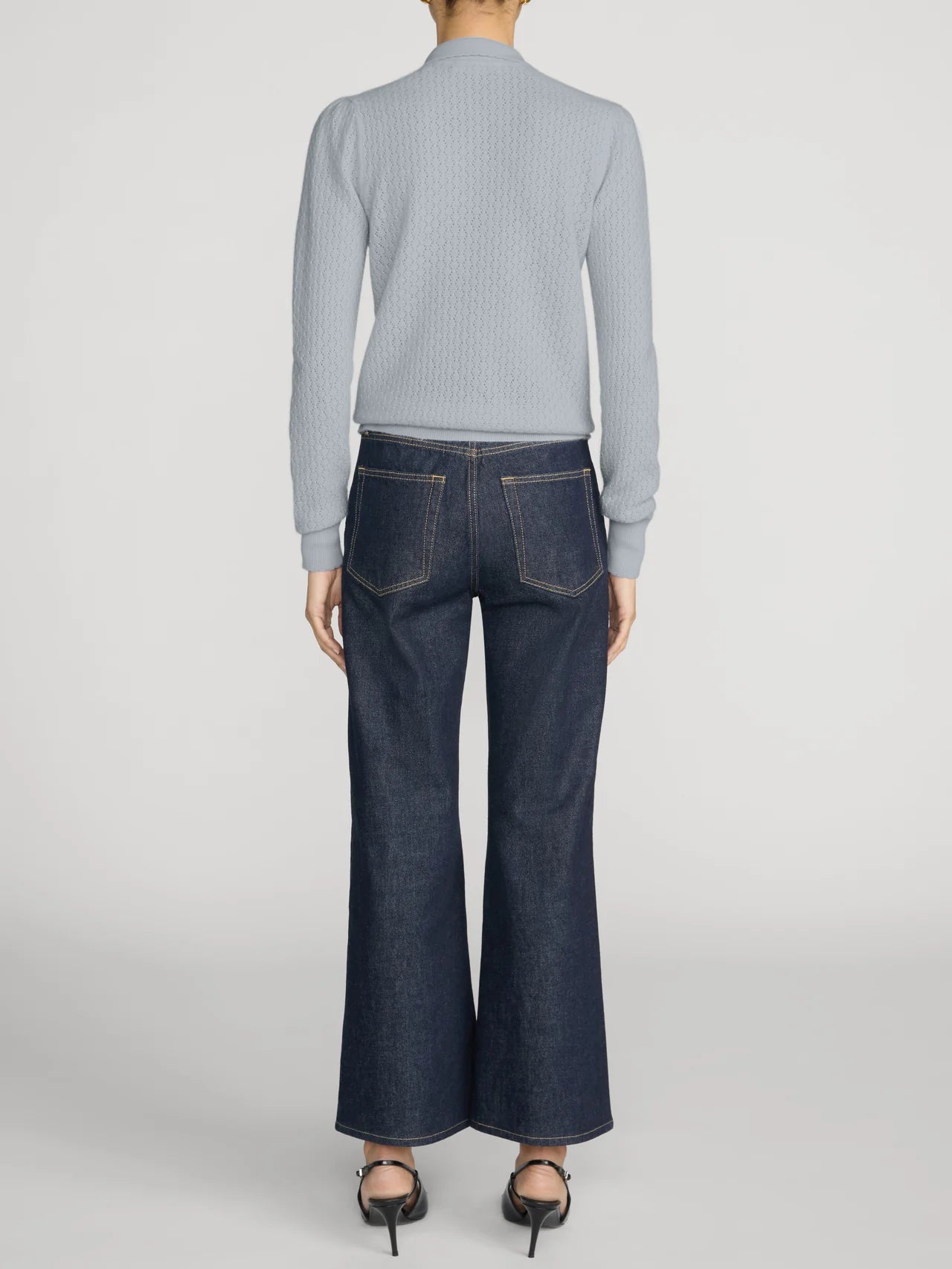 A model shown from the back wearing a light gray ribbed mock-neck sweater and dark blue wide-leg denim jeans, paired with black high heels, against a light gray studio background.